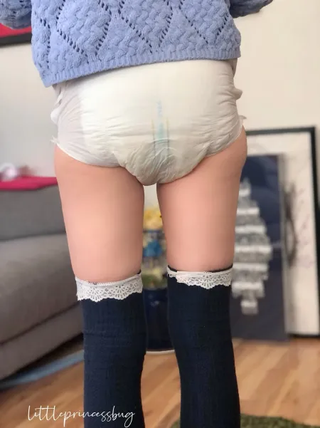 A cute girl from behind in thick white adult diaper and navy lace-top thigh-high socks.