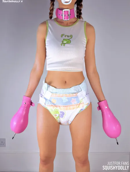 A girl in a pacifier gag and pigtails wearing a colorful printed diaper and pink inflatable gloves