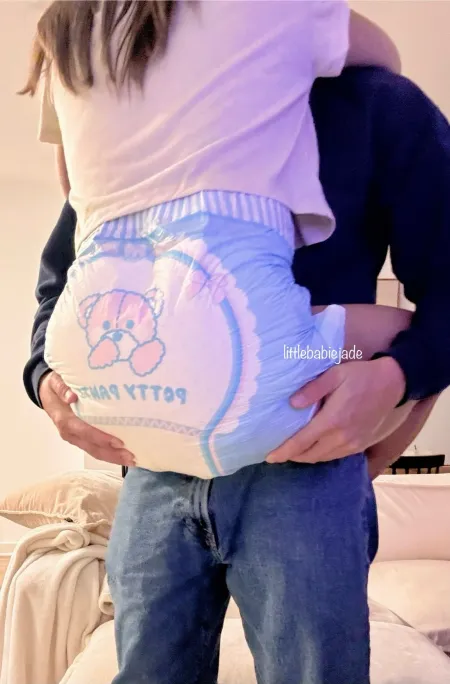 A girl being held up by someone, her huge bulging diaper with teddy bear print fully exposed from behind.