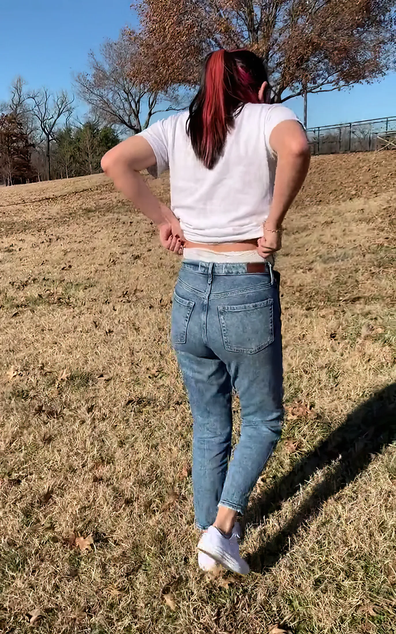 An ABDL girl adjusts the waistband of her diaper peeking out over her jeans while outdoors.