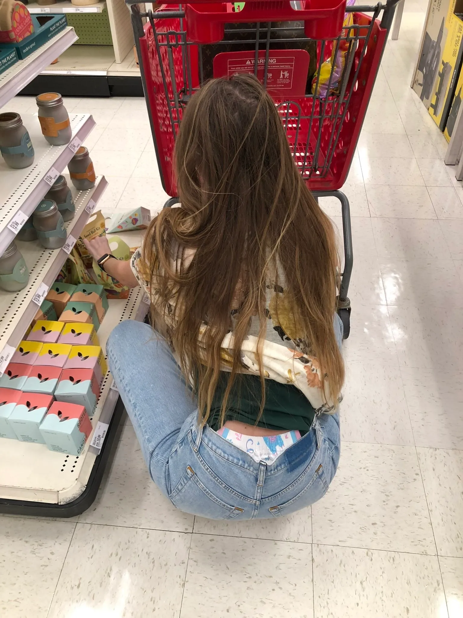An ABDL girl squats down at the supermarket, exposing her diaper waistband.