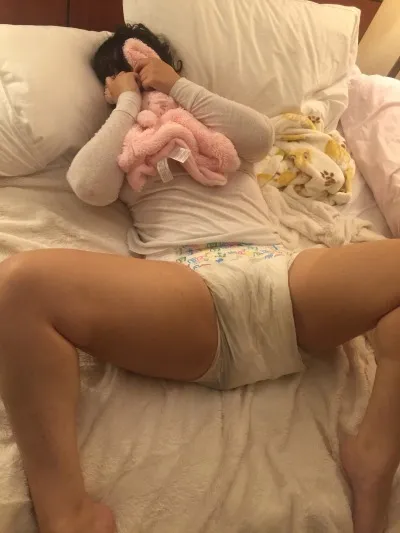 An ABDL girl lies in bed, covering her face and spreading her legs to show off her wet diaper.