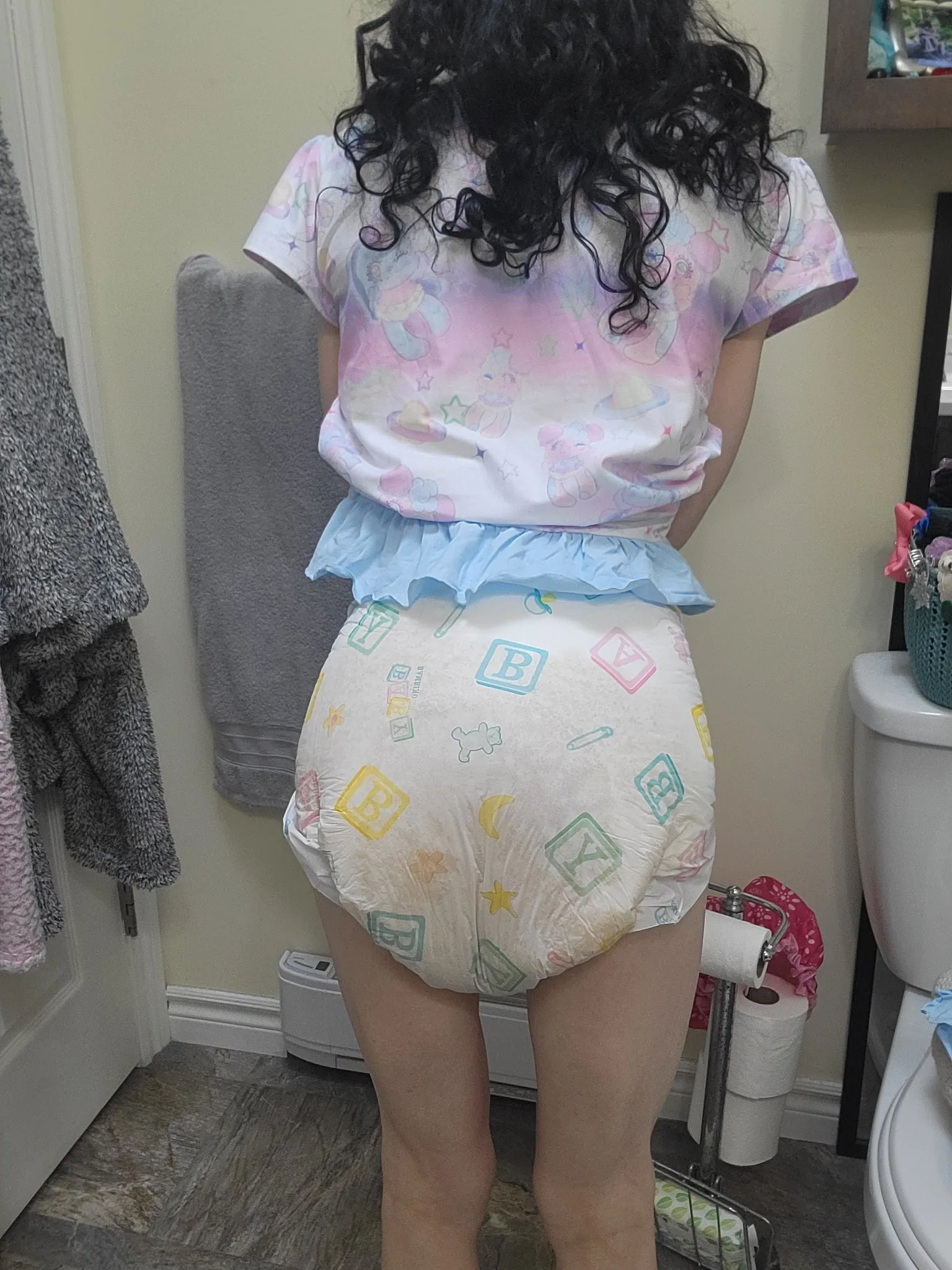 ABDL girl flaunts diaper silhouette beneath red undershirt.