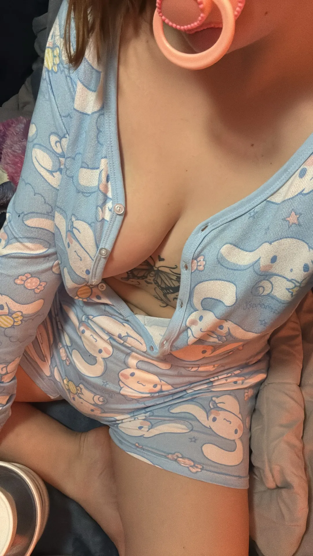 A girl biting a pacifier, her blue romper buttons undone to reveal part of her breast.