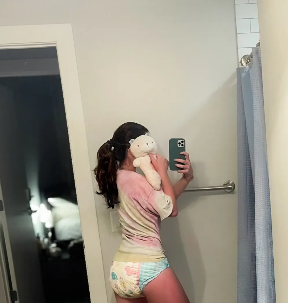 A pretty girl holding a teddy bear takes a mirror selfie, wearing a cute top with pastel-colored soiled ABDL diapers.