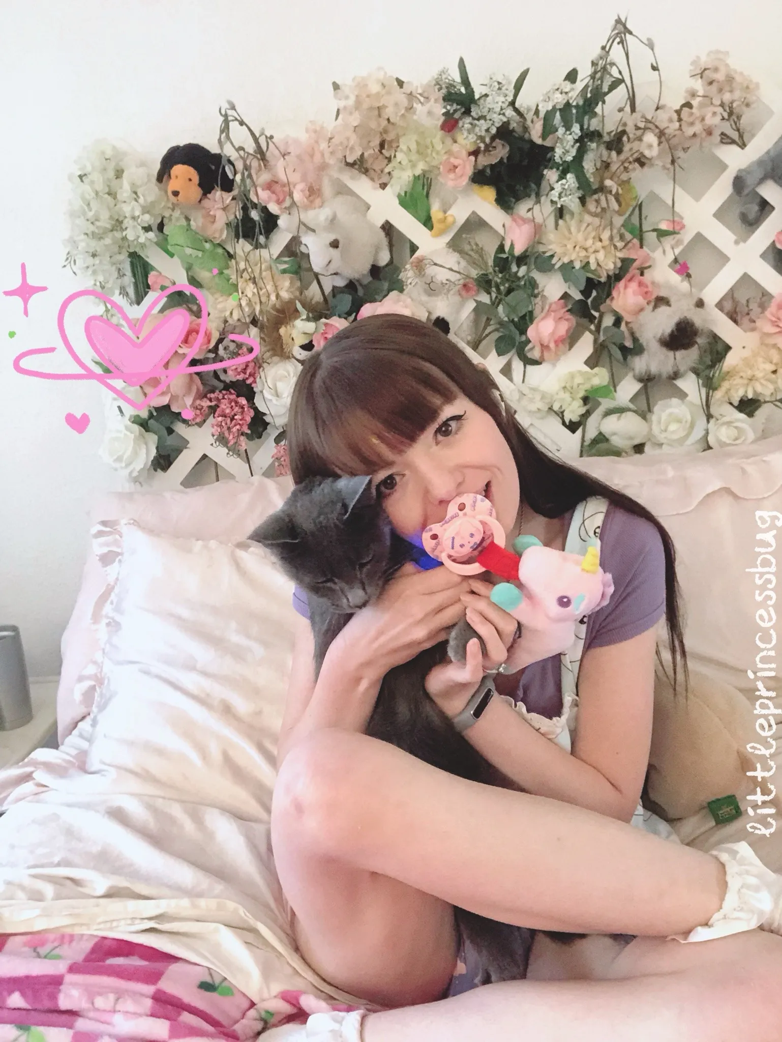 A cute girl sucking on a lollipop in front of a flower wall, hugging a black cat, with her thighs and purple diaper exposed.