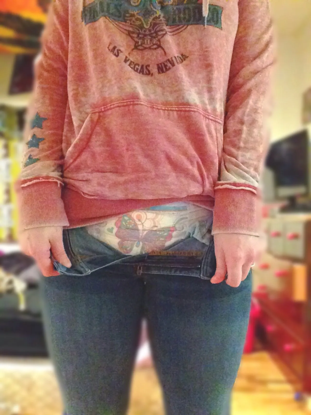 A young woman wearing a Vegas hoodie has her jeans pulled down low, clearly revealing her cute patterned diaper.